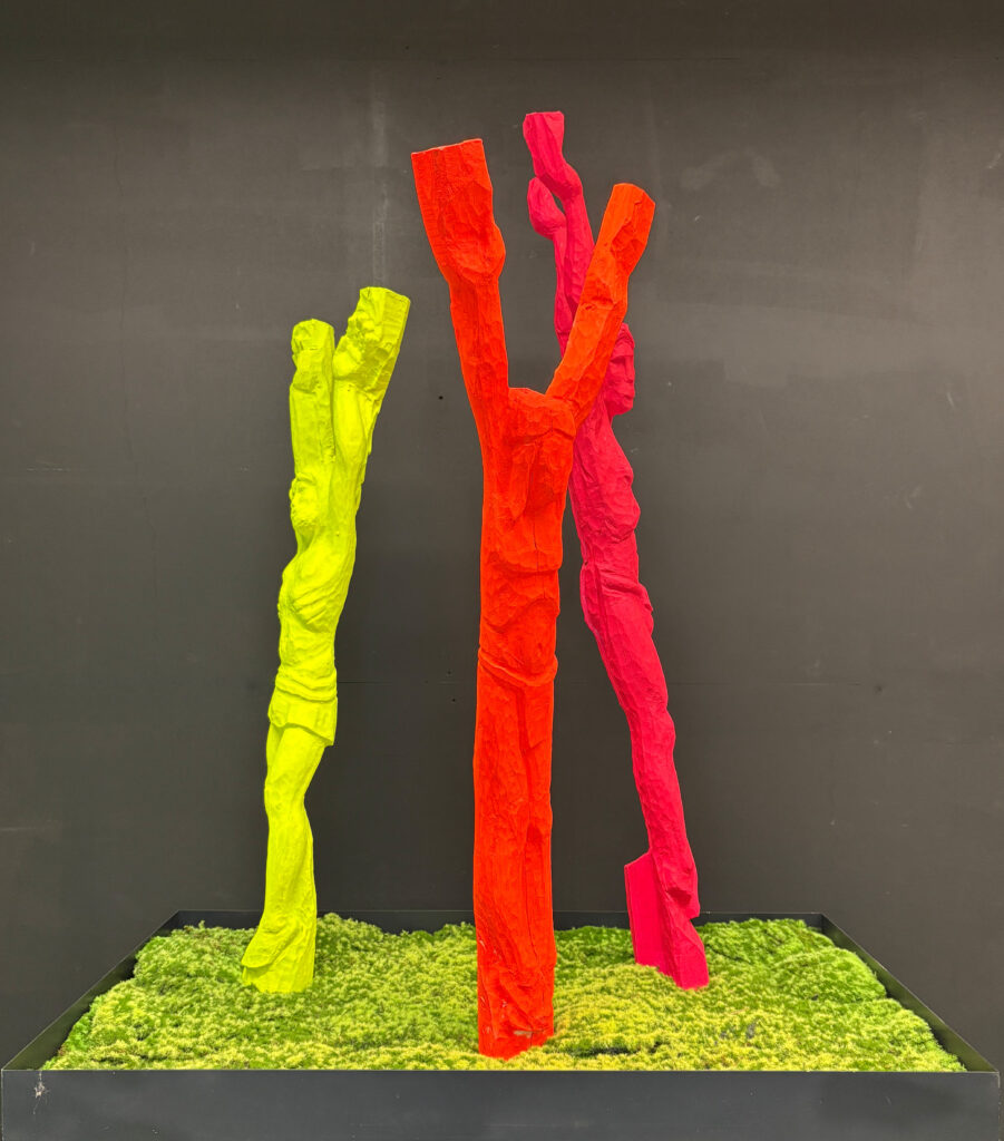 Auf dem Bild sind vier neonfarbene Skulpturen, die Neon Gods, zu sehen, die in einer schwarzen Pflanzschale stehen, die mit Moos ausgekleidet ist. Die linke Figur ist neongelb, die mittlere Figure ist neonoorange, die rechte Figur ist neonpink. Die Oberflächen der Figuren wirken stark strukturiert oder schichtartig modeliert, was ihnen eine organische Anmutung verleiht.